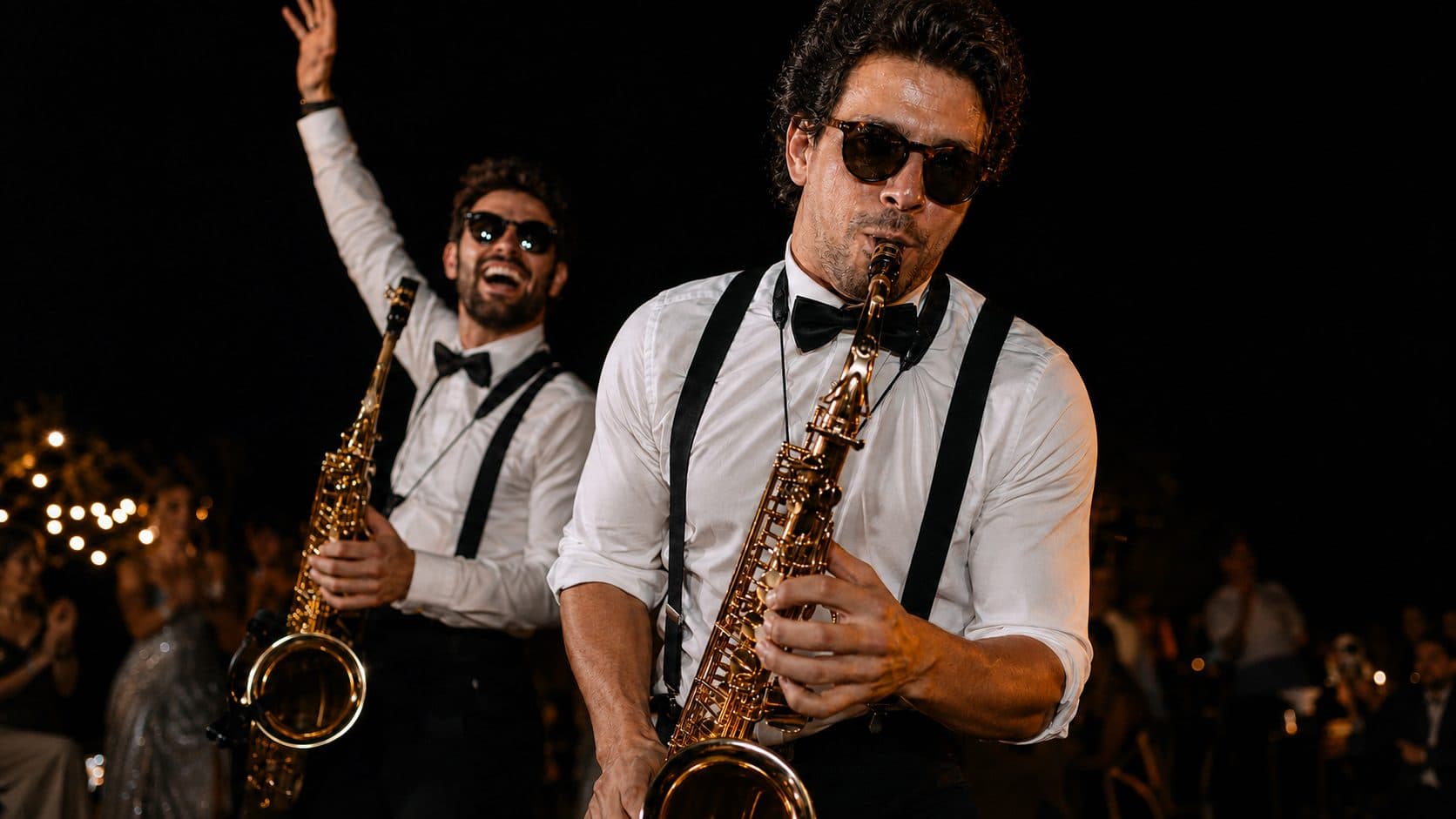 Deux saxophonistes en bretelles et lunettes noires jouent au milieu des invités, la nuit