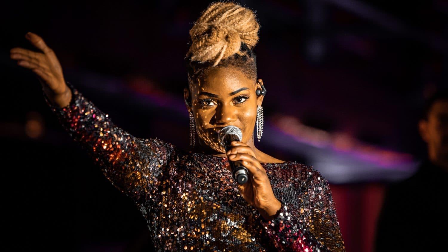 Chanteuse en veste à sequins au micro, bras levé, sous une lumière violette