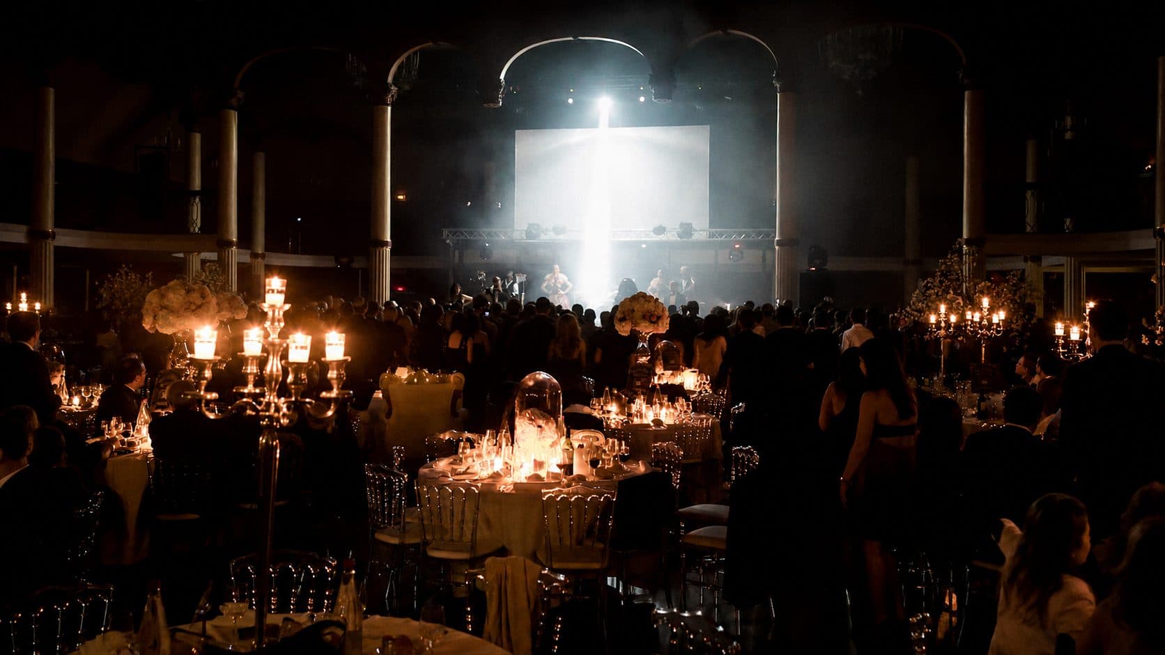 Salle de gala aux chandeliers, tables dressées et écran géant, invités debout devant la scène