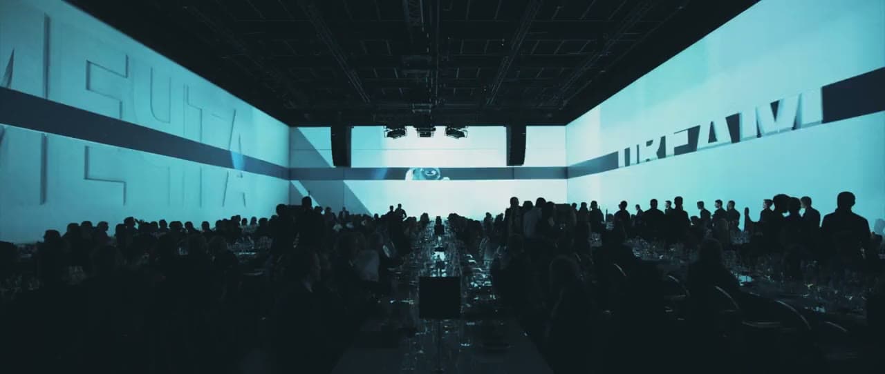 Public en silhouette dans un auditorium sombre, face à un écran LED géant bleu turquoise, lors d’un événement d’entreprise