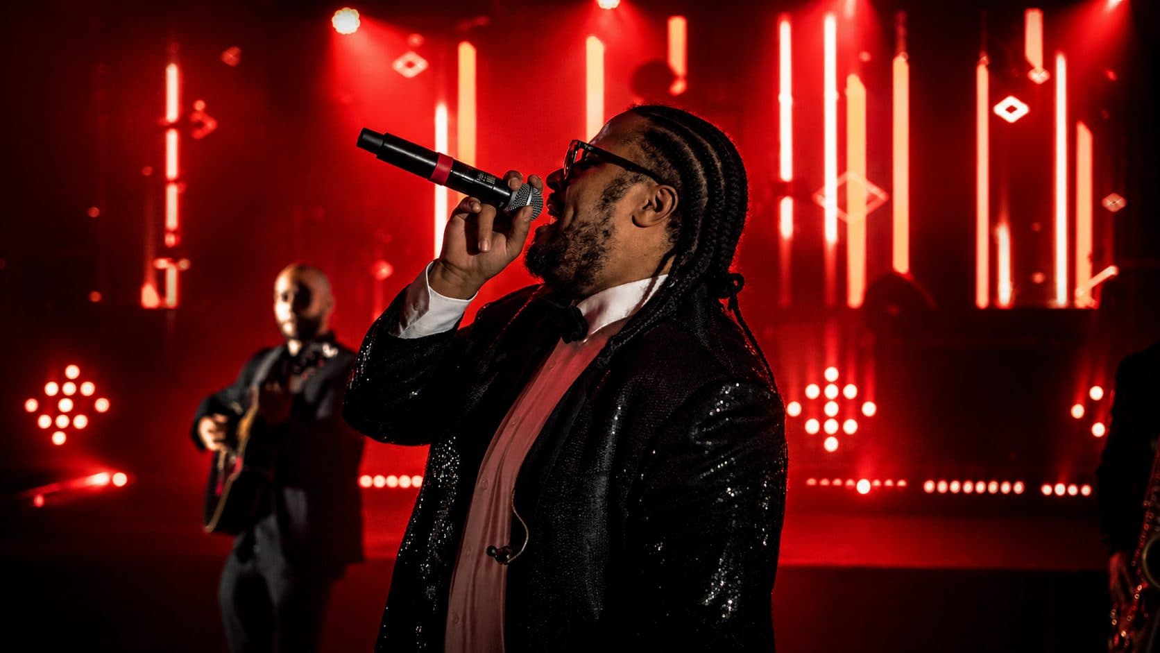 Chanteur au micro en veste à paillettes devant un décor de néons rouges, guitariste en retrait
