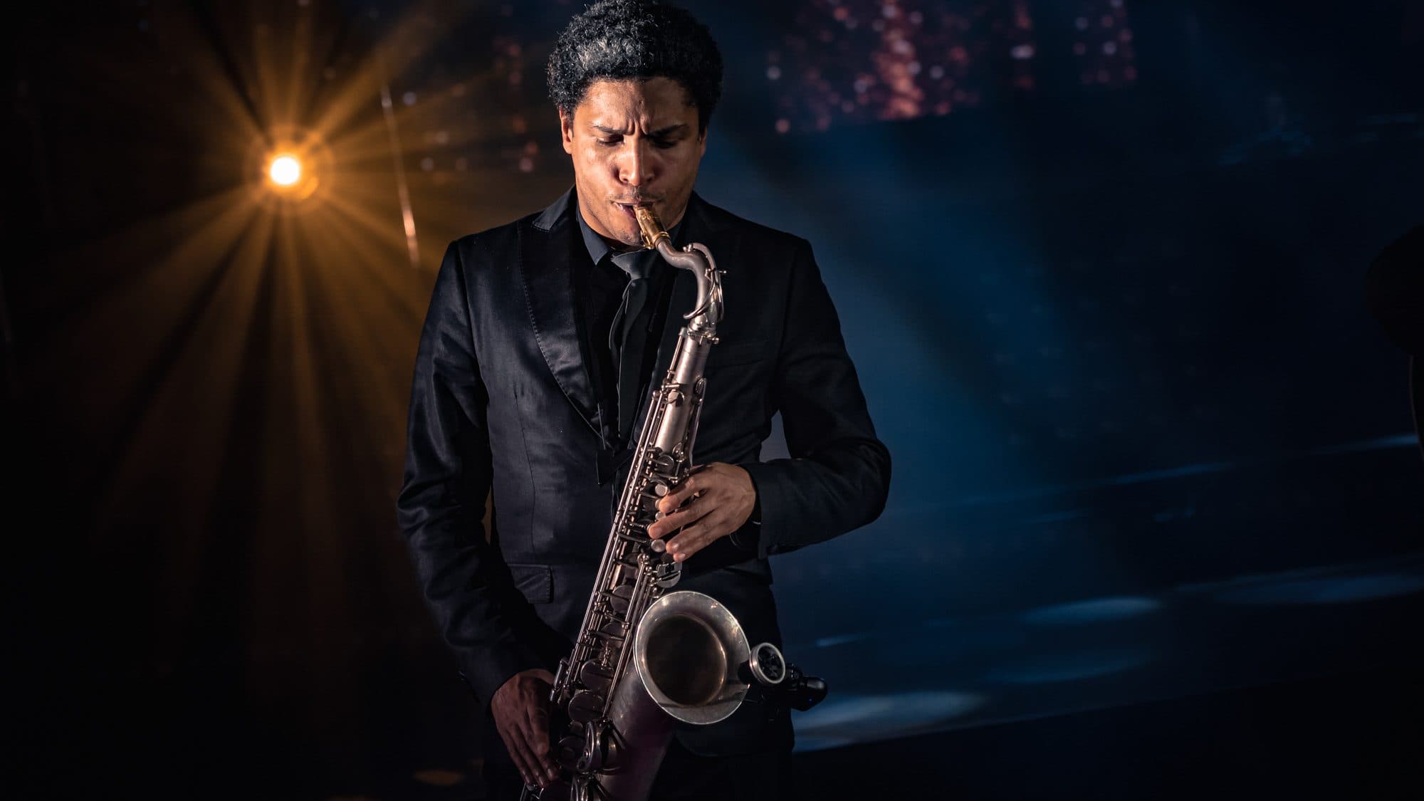 Saxophoniste en costume noir jouant sur scène, à contre-jour d’un projecteur ambré