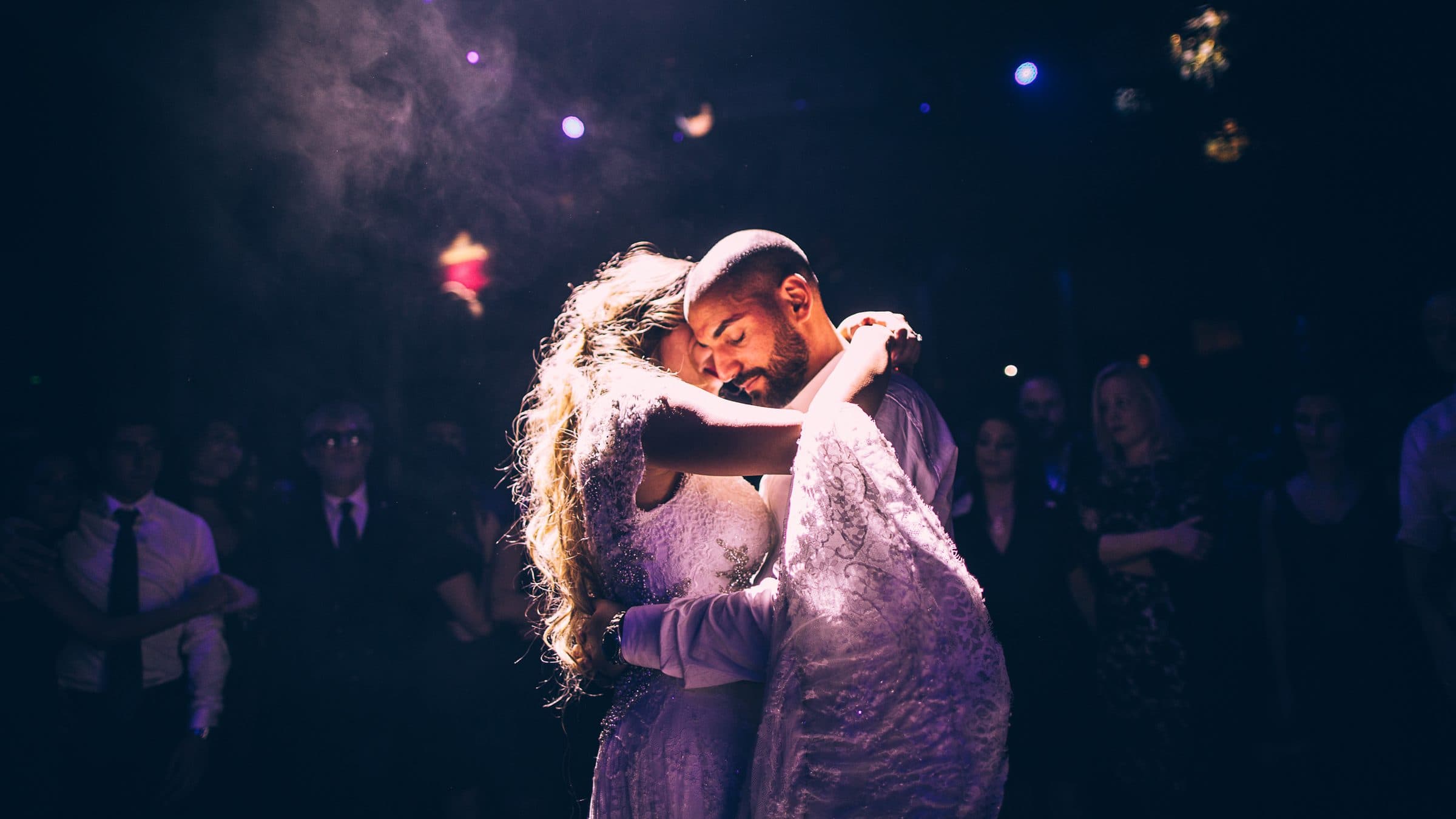 Mariés enlacés pendant leur première danse, sous une lumière violette, entourés de leurs invités
